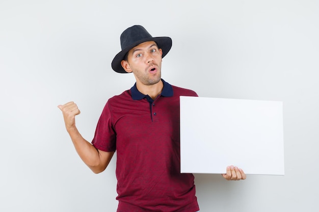 Giovane uomo in t-shirt, cappello che punta il pollice indietro, tiene in mano tela e sembra meravigliato, vista frontale.