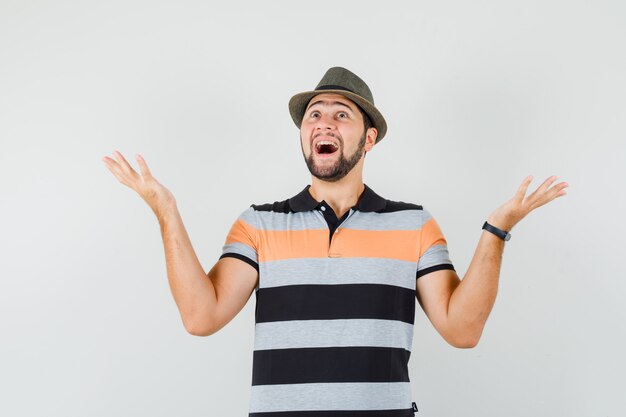 Молодой человек в футболке, шляпе, поднимая руки, глядя вверх и глядя весело, вид спереди.