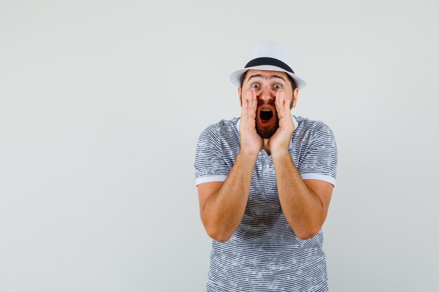 Young man in t-shirt, hat shouting or announcing something and looking anxious , front view.