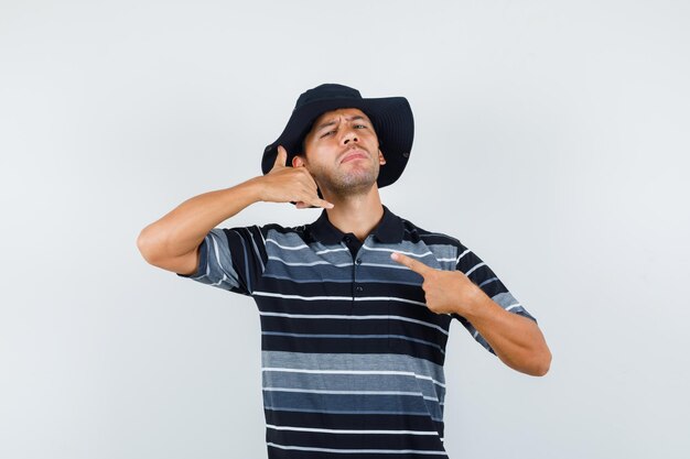 Young man in t-shirt, hat showing call me gesture and looking confident , front view.
