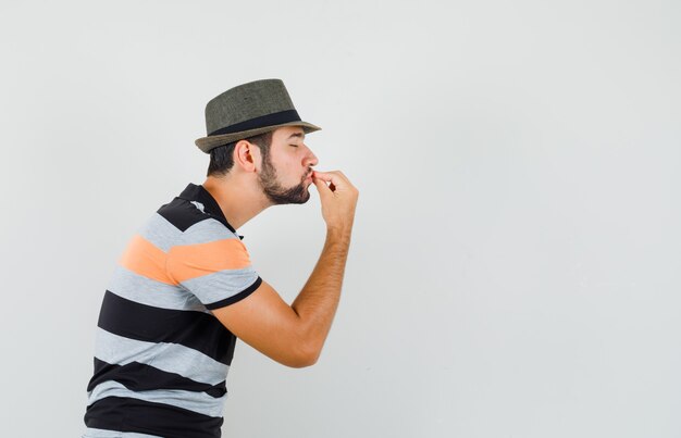 Tシャツを着た若い男、指にキスをし、喜んで見えることでおいしいジェスチャーを示す帽子。