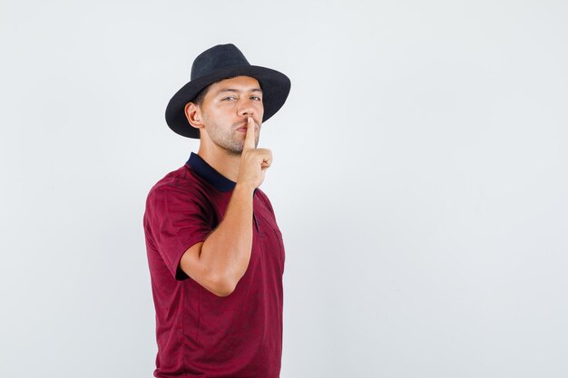 Young man in t-shirt, hat showing silence sign and looking careful , front view.