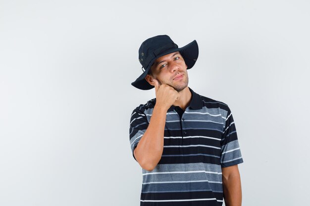 Tシャツを着た若い男、頬の近くに親指を表示し、陽気に見える帽子、正面図。