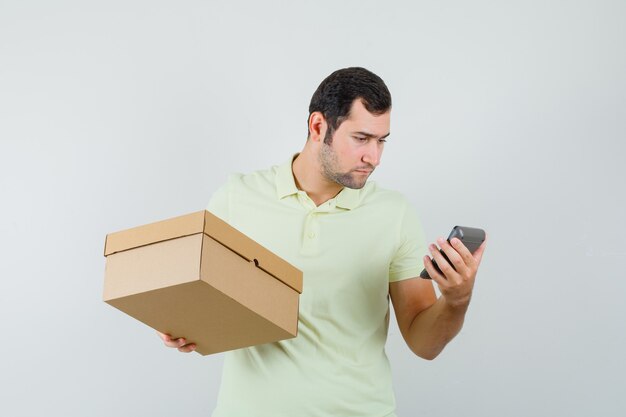 Young man in t-shirt holding cardboard box, looking at calculator , front view.