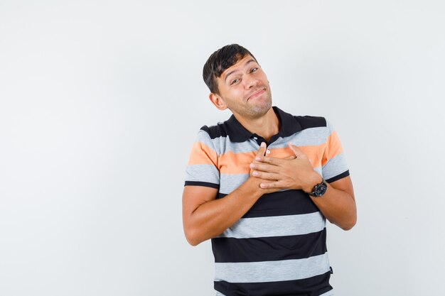 Young man in t-shirt holding hands on chest and looking cheerful