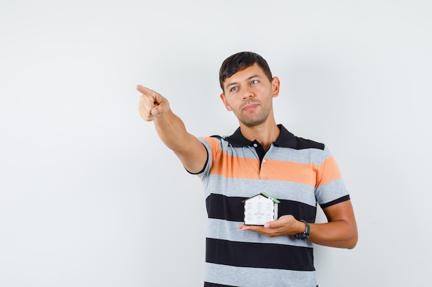Young man in t-shirt holding house model while pointing away and looking focused 