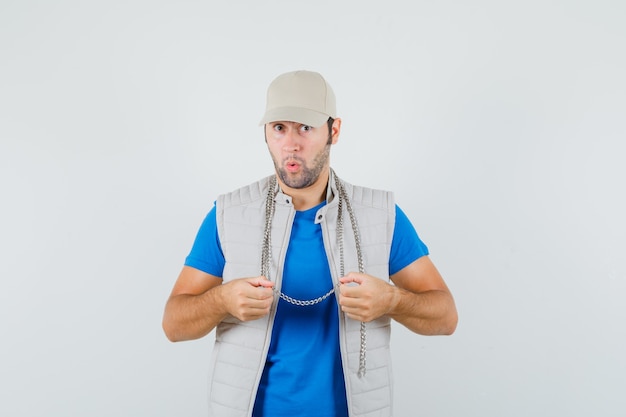 Young man in t-shirt, jacket, cap holding his chain necklace and looking amazed , front view.