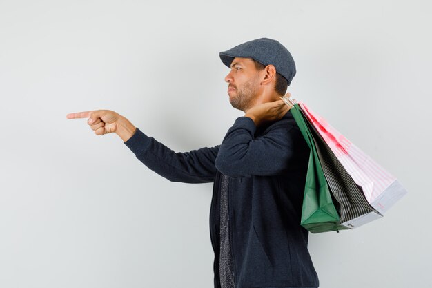 Young man in t-shirt, jacket, cap holding shopping bags, pointing to the front of him .