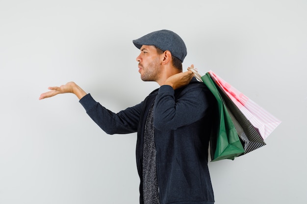 Free photo young man in t-shirt, jacket, cap holding shopping bags, sending air kiss .