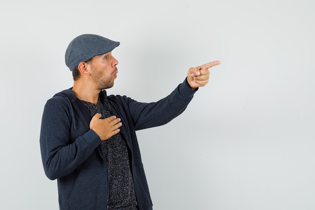 Free photo young man in t-shirt, jacket, cap pointing away, holding hand on chest and looking amazed , front view.