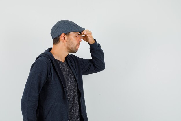 Young man in t-shirt, jacket, cap rubbing eyes and nose and looking fatigued , front view.