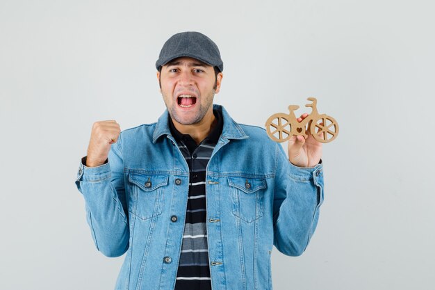 Young man in t-shirt, jacket, cap showing winner gesture, holding wooden toy bike and looking lucky