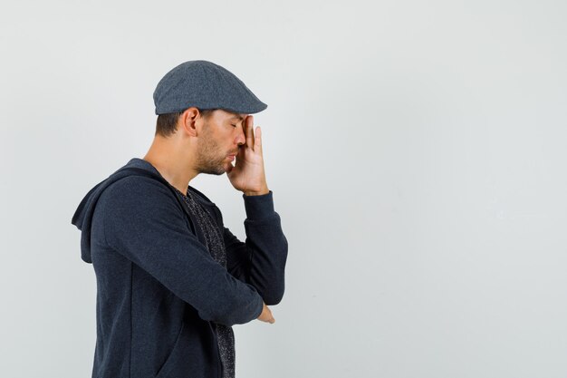 Young man in t-shirt, jacket, cap standing in thinking pose with closed eyes and looking tired .