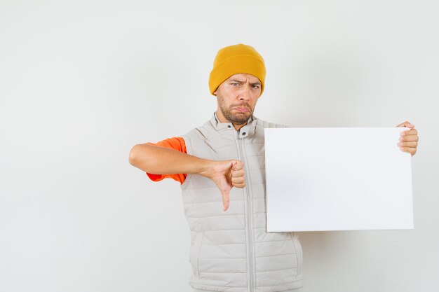 Tシャツ、ジャケット、空白のキャンバスを保持している帽子、親指を下に表示し、失望しているように見える、正面図の若い男。