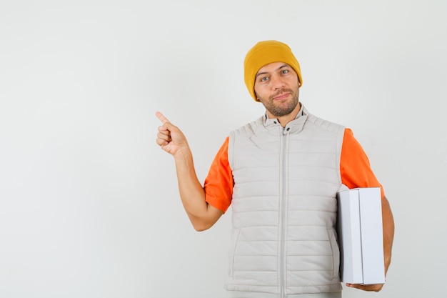 Young man in t-shirt, jacket, hat pointing away, holding cardboard box