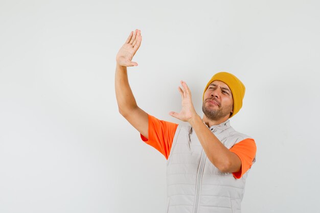 Молодой человек в футболке, куртке, шляпе поднимает руки, чтобы защитить себя и выглядит испуганным