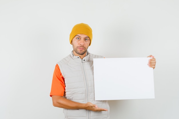 Young man in t-shirt, jacket, hat showing blank canvas and looking positive