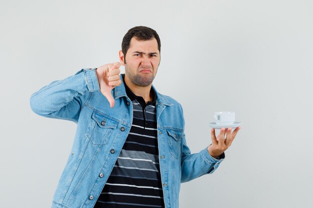 Молодой человек в футболке, пиджаке держит чашку чая, показывает палец вниз и выглядит недовольным