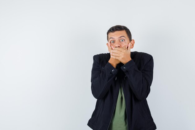 Young man in t-shirt, jacket holding hands on mouth and looking scared , front view.