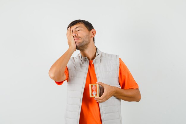 Young man in t-shirt, jacket holding hourglass with hand on face and looking forgetful , front view.