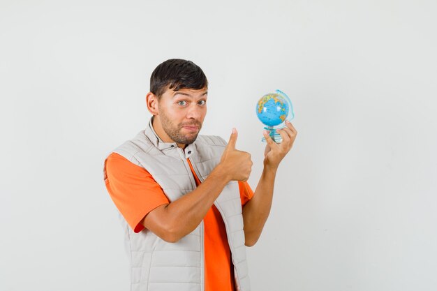 young man in t-shirt, jacket holding school globe, showing thumb up and looking cheery , front view.