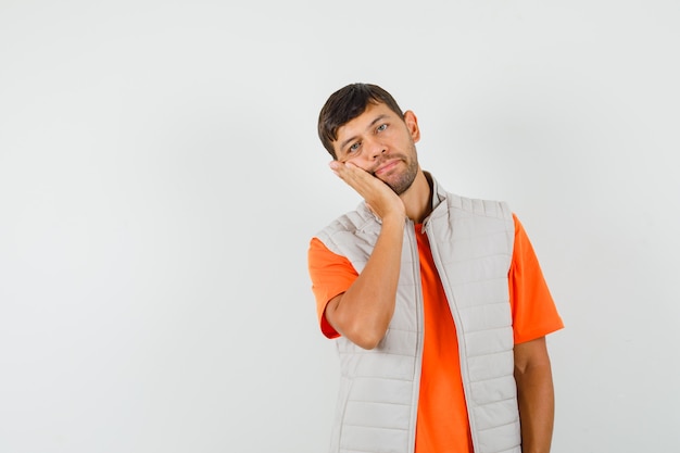 Young man in t-shirt, jacket leaning cheek on raised palm and looking pensive , front view.