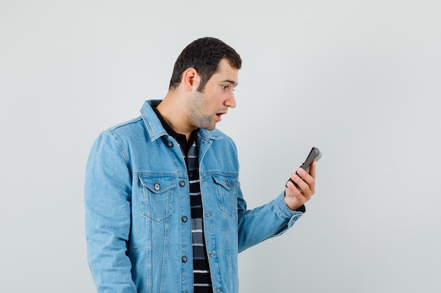 Young man in t-shirt, jacket looking at calculator and looking wondered