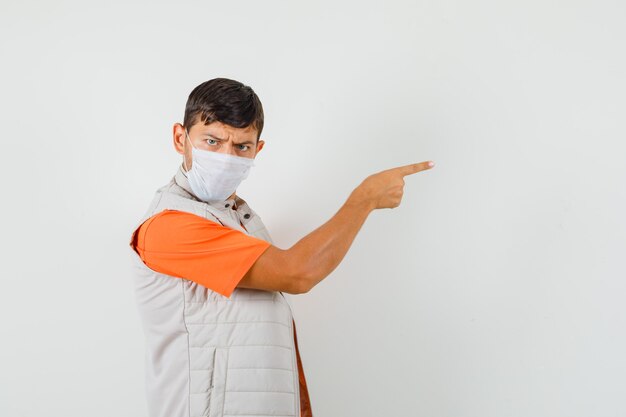 Young man in t-shirt, jacket, mask pointing to the side and looking serious .