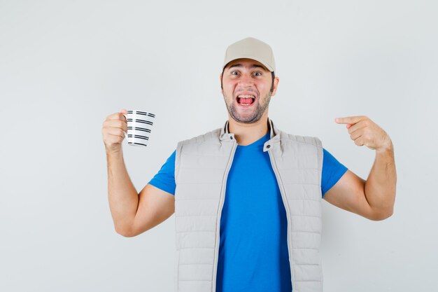 Young man in t-shirt, jacket pointing at cup of drink and looking confident , front view.