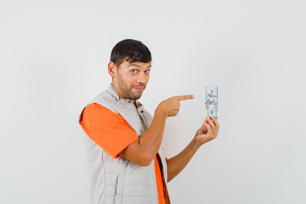 Young man in t-shirt, jacket pointing at dollar bill and looking cheery , front view.