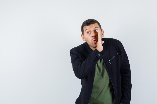 Young man in t-shirt, jacket telling secret behind hand and looking curious , front view.