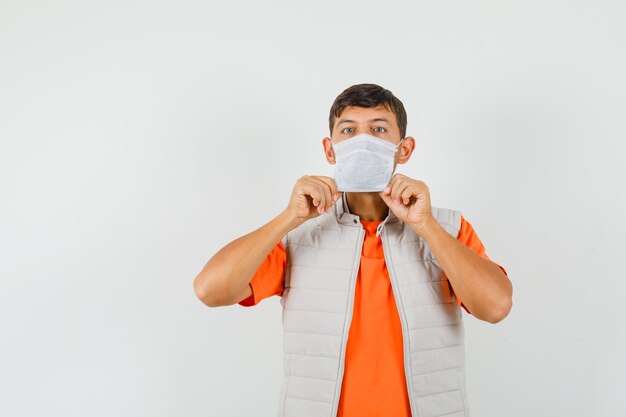 Young man in t-shirt, jacket wearing medical mask and looking careful , front view.