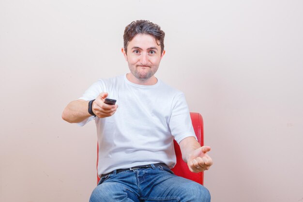 Young man in t-shirt, jeans using remote controller while sitting on chair