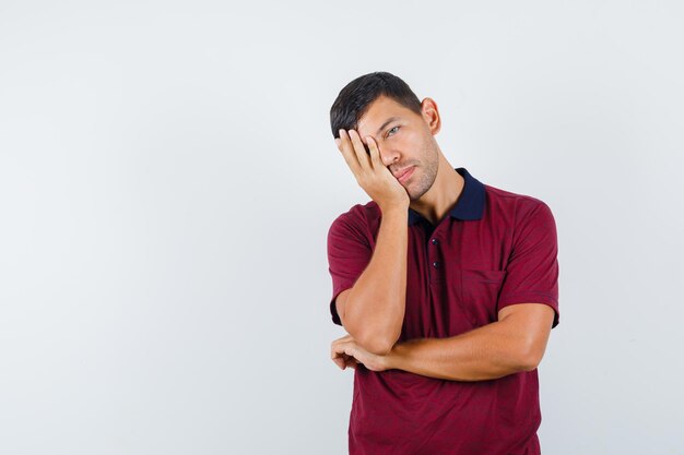 Young man in t-shirt leaning head on raised palm and looking tired , front view.