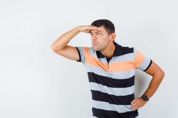 Young man in t-shirt looking far away with hand over head and looking focused 