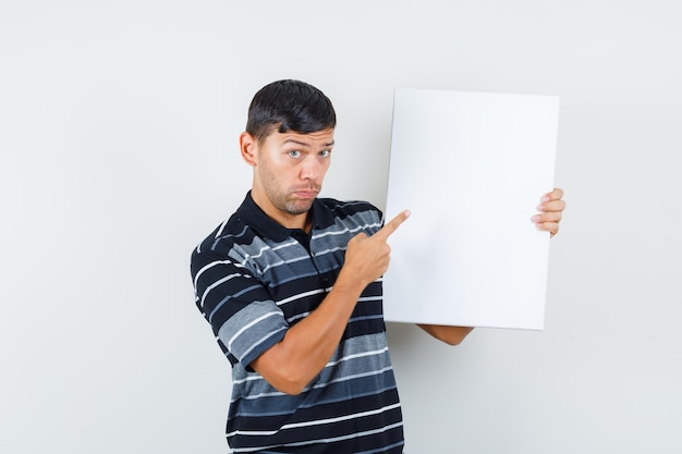 Young man in t-shirt pointing at blank canvas and looking hesitant , front view.