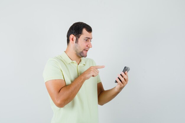 Young man in t-shirt pointing at calculator and looking confused, front view.