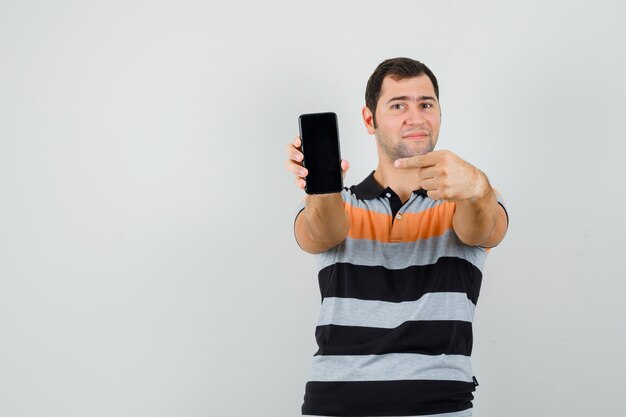 Молодой человек в футболке указывает на мобильный телефон и выглядит довольным