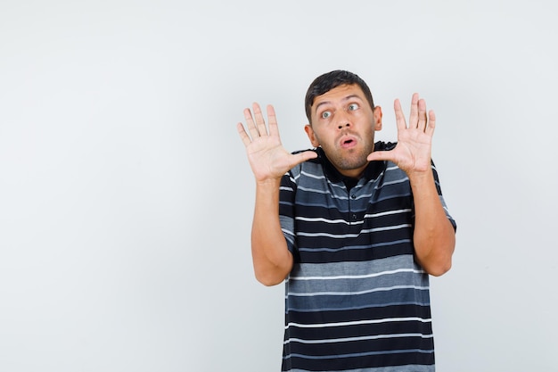 Young man in t-shirt raising hands in preventive manner and looking scared , front view.