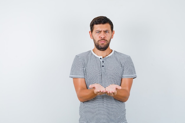 Young man in t-shirt showing receiving or giving gesture and looking gloomy , front view.