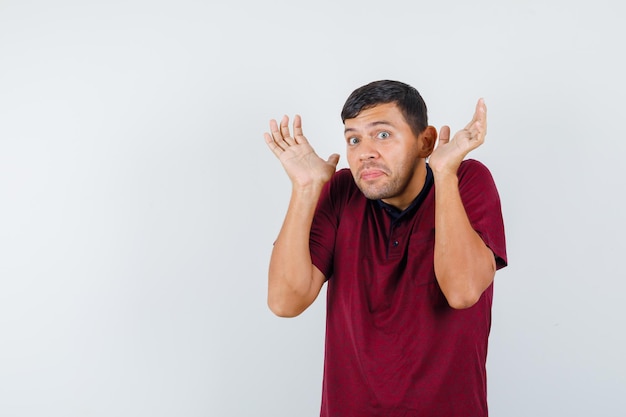 Young man in t-shirt shrugging, raising hands in surrender gesture and looking scared , front view.