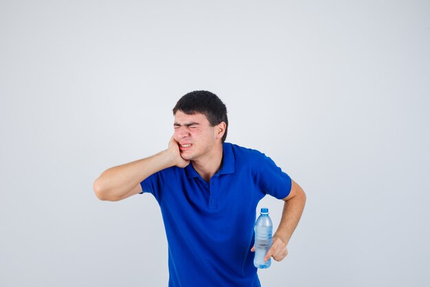 Молодой человек в футболке страдает от зубной боли, держит пластиковую бутылку и выглядит болезненно, вид спереди.