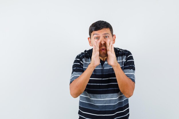 Young man in t-shirt telling secret by holding hands near mouth , front view.