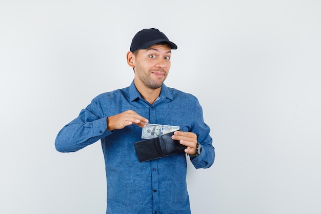 Free photo young man taking dollar bill out of wallet in blue shirt, cap and looking joyful. front view.