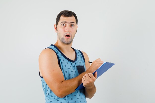 Young man taking notes on clipboard in blue singlet and looking amazed , front view.