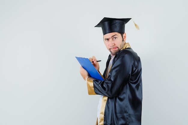 Young man taking notes on clipboard in graduate uniform and looking doubtful .