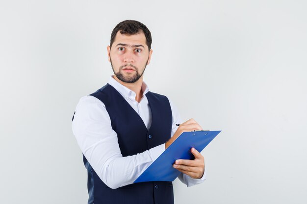 Young man taking notes on clipboard in shirt and vest and looking focused