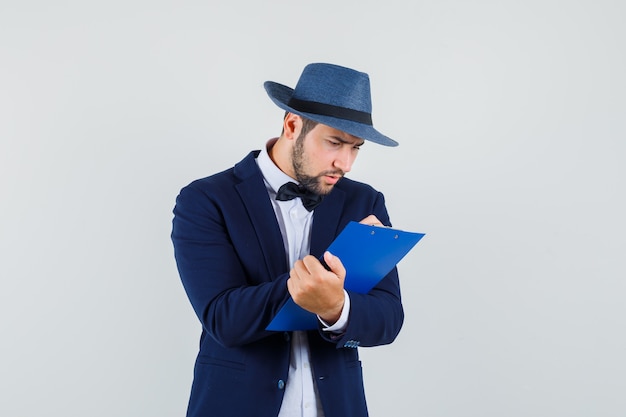 Giovane che prende appunti sugli appunti in vestito, cappello e che sembra occupato. vista frontale.