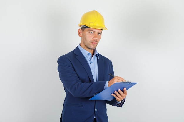 Young man taking notes on clipboard in suit, safety helmet and looking busy