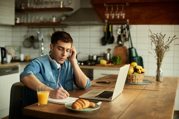 Young man taking notes while listening online class over laptop at home